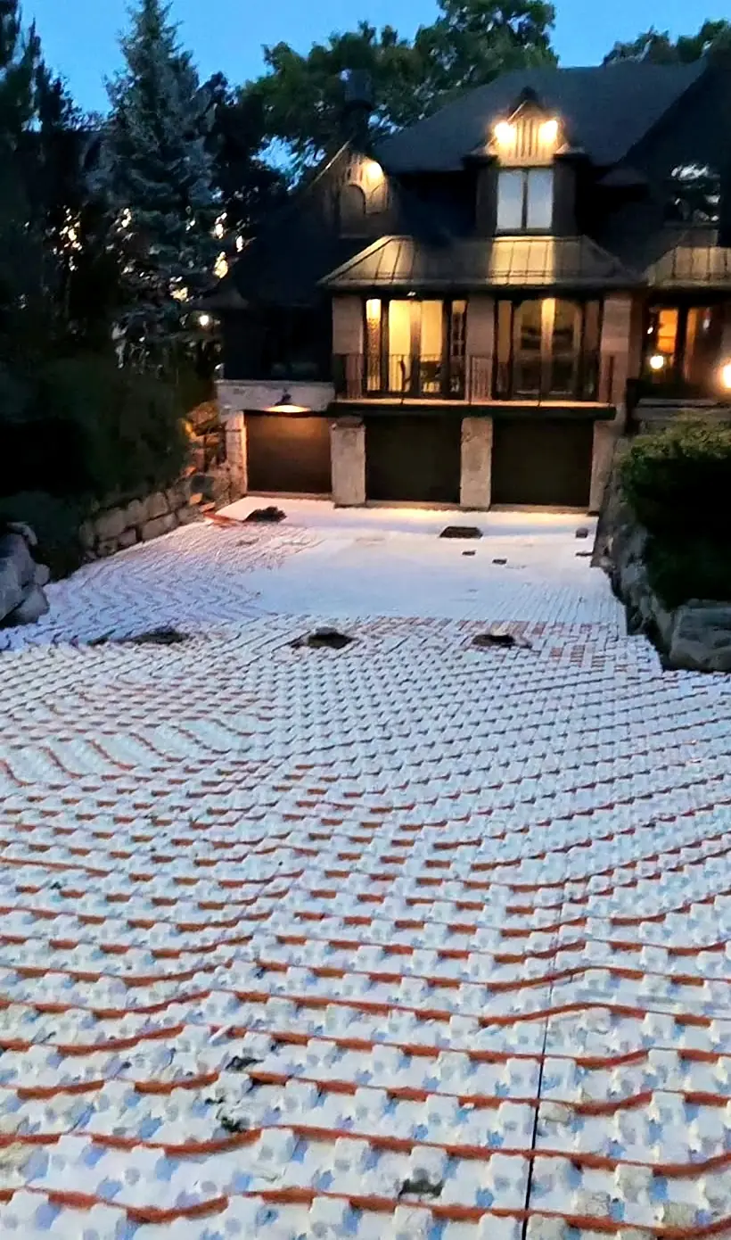 A residential driveway covered in snowmelt heating loops at dusk, with the house lit behind
